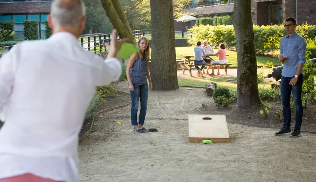 Personen spielen Cornhole im Garten mit Holzbrett und Beanbags.